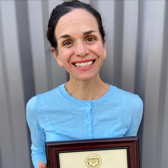 Ana Pinto da Silva, brown hair, brown eyes, smiling broadly and holding a framed certificate of recognition from the County of Orange.