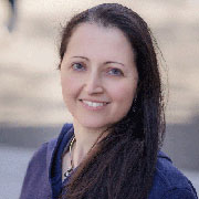 Anat Caspi: A white woman smiling into the camera. She is wearing a purple blouse.