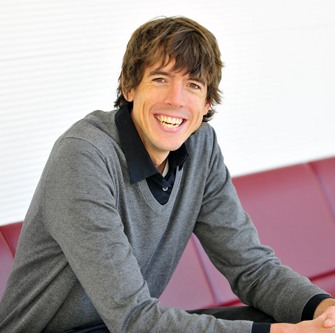 Jon Froehlich. He is smiling and sitting on a red couch. He is wearing a grey shirt.