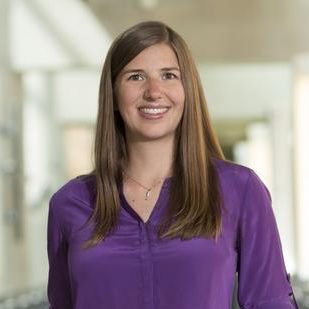 Kat Steele, standing on a bridge and wearing a purple blouse. Her hair is down and she is smiling