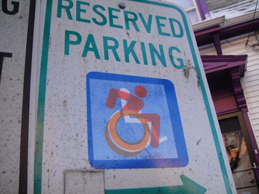 The revised disabled symbol, a person with their arms back to go quickly in a manual wheelchair, in red shown on a parking sign over the old symbol.