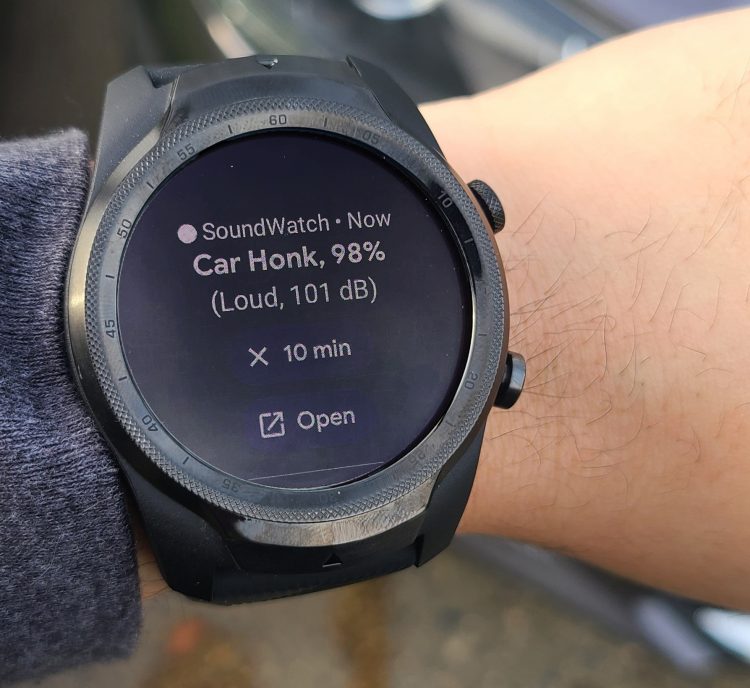 A wrist with a smartwatch on it. The smartwatch has an alert that says "Car honk, 98%, Loud, 101 dB" It also has options to snooze the alert for 10 minutes or open in an app on the user's phone.