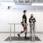 Researcher Alyssa Spomer uses a tablet to monitor a man wearing a robotic exoskeleton device around his hips and legs and walking on a treadmill.