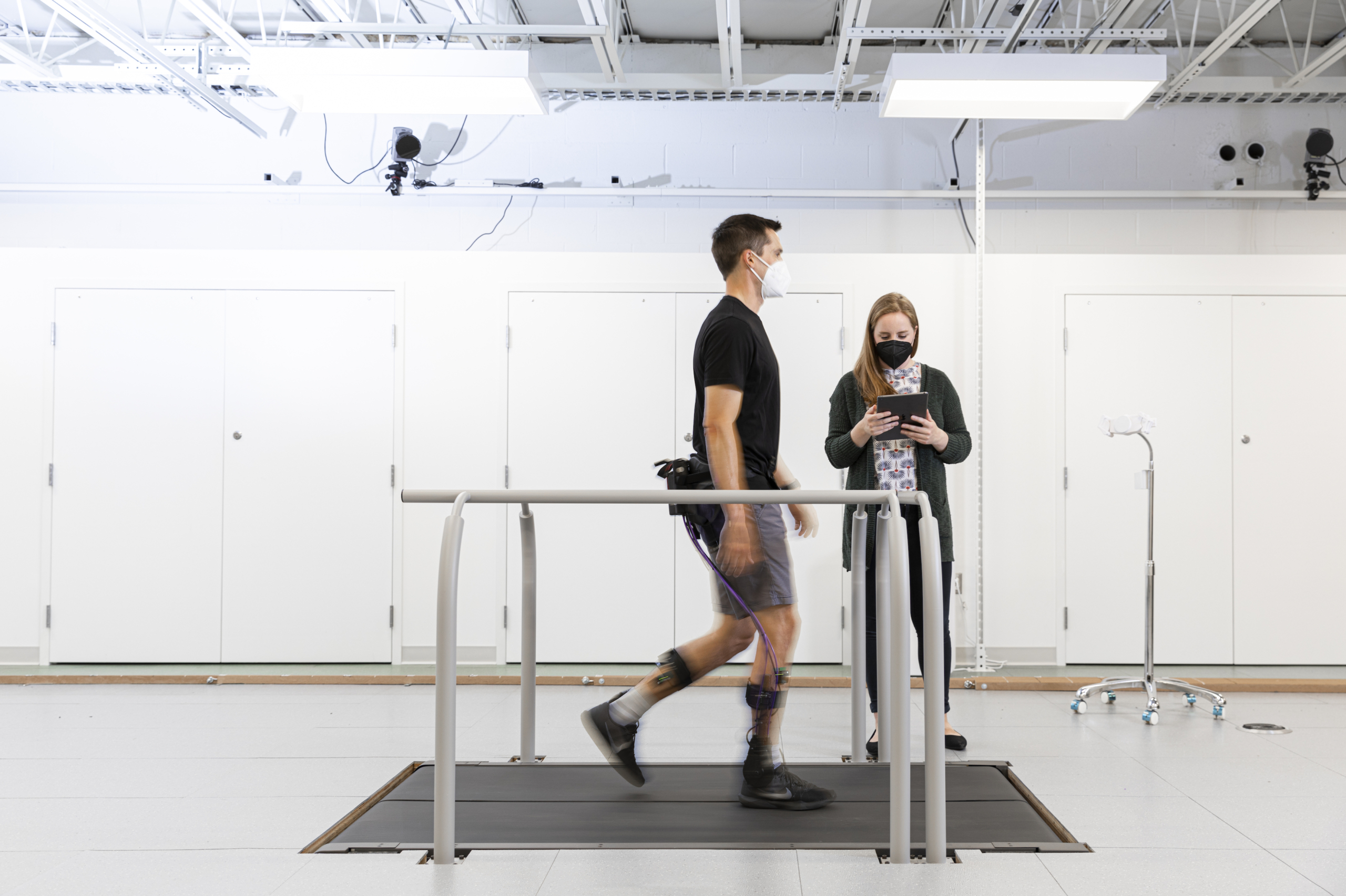 Researcher Alyssa Spomer uses a tablet to monitor a man wearing a robotic exoskeleton device around his hips and legs and walking on a treadmill.