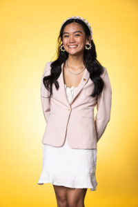 Kianna Bolante stands in a white dress and pink blazer in front of a vivid gold background for the Husky100 photo series.