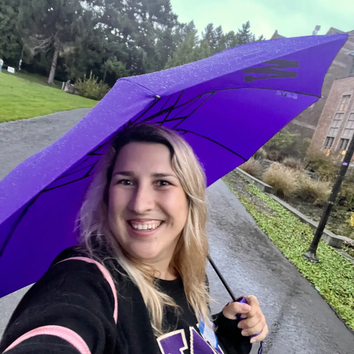 Kate Glazko, a white woman in a UW sweatshirt grinning while holding a purple umbrella in the rain.