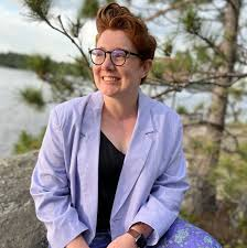 Emma McDonnell, seated in front of pine trees and distant water. She is wearing a light purple blazer.