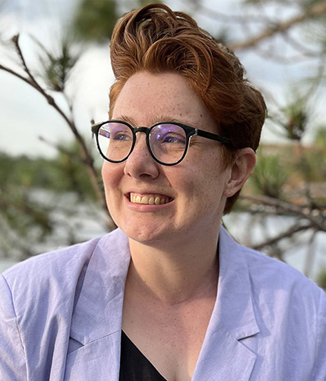 Headshot of EmmaMcDonnell, with pine trees and distant water in the background. She is wearing a light purple blazer and gazing into the distance.