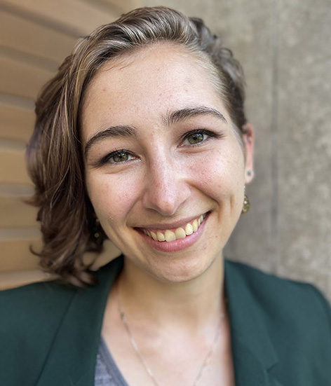 Kelly Avery Mack, a white, femme-presenting person with curly light brown hair shaved close on one side wearing a green blazer and grey top.