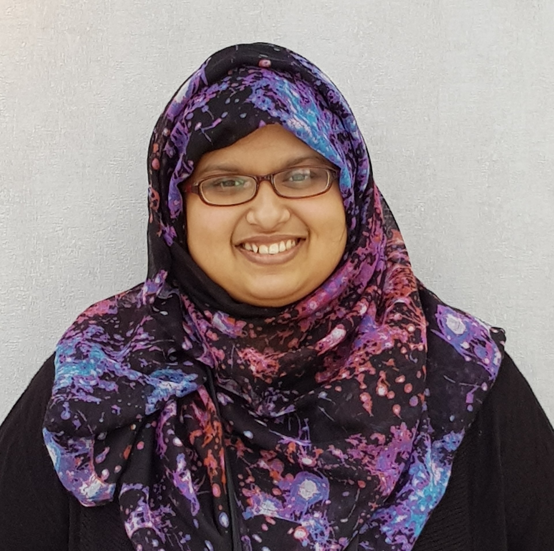 Headshot of Tamanna Motohar. She is wearing glasses and a colorful hijab, smiling at the camera, standing against a plain light gray background