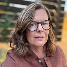 Headshot of Liz Diether-Martin, a white 50+ woman wearing brown glasses, a brown jacket and standing in front of a garden fence