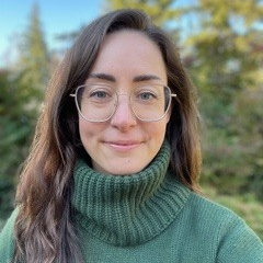 Headshot of Bethany Sloane. Bethany has long, brown hair, gold glasses, and is wearing a green sweater. She is outside with pine tree’s and a blue sky in the background.