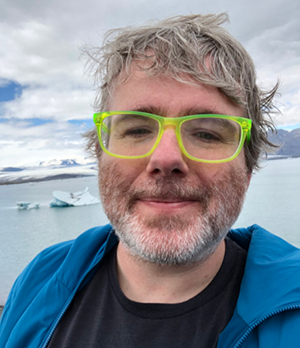 Shaun has grey hair and a grey beard and bright green glasses. In this image he is posing in front of a frozen lake in Iceland.