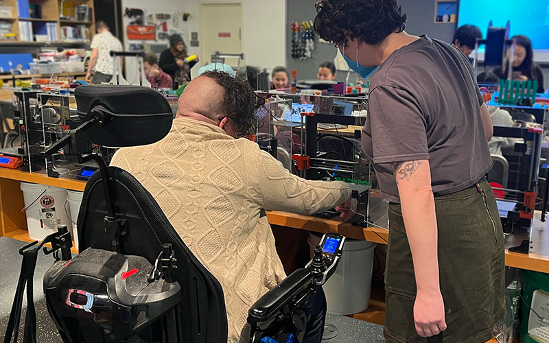 At a 3D printer, two adaptation hackfest participants checking progress. One person is using a power wheelchair and the other is standing.