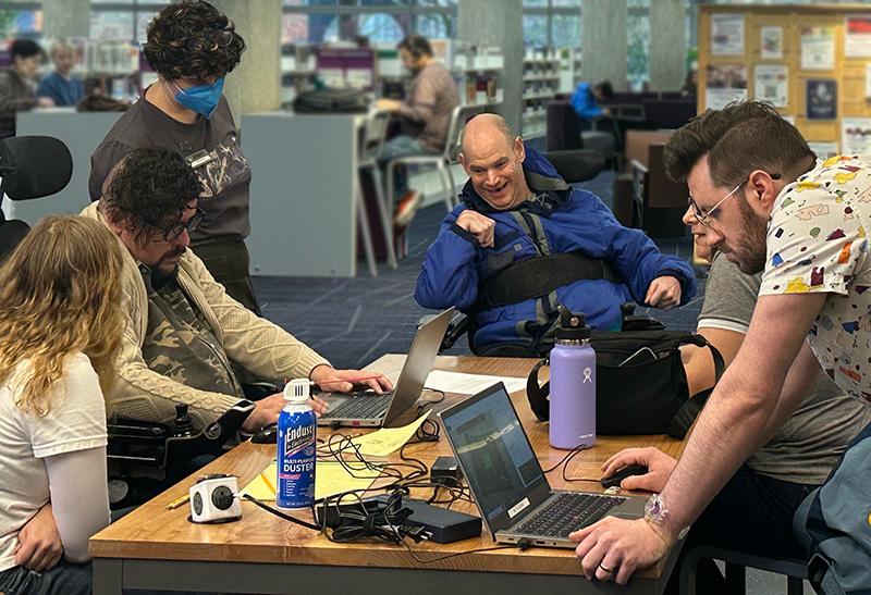 A group of 5 adaptation hackfest participants in discussion at a table in the library. One is using a power wheelchair and two are working on laptops. 