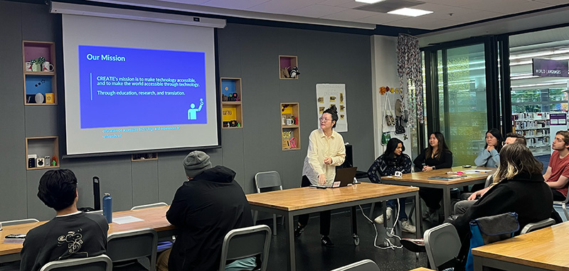 An adaptation hackfest participant presents their team's challenge. Behind them is an overhead with their challenge: to create a prototype portable bidet.