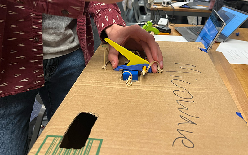 Closeup of an adaptation hackfest prototype for a microwave button pressing tool. A cardboard box is used to represent the microwave and a 3D printed  lever is placed on top.