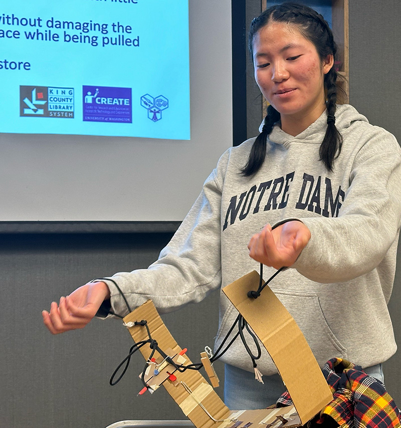 An adaptation hackfest participant presents their team's prototype: A device to assist with pulling up pants for a person who uses prosthetics. They have maneuvered their arms into loops attached to a cardboard belt.