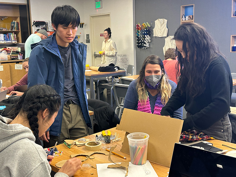 Four adaptation hackfest participants working at a table covered in cardboard, tools, and a laptop.