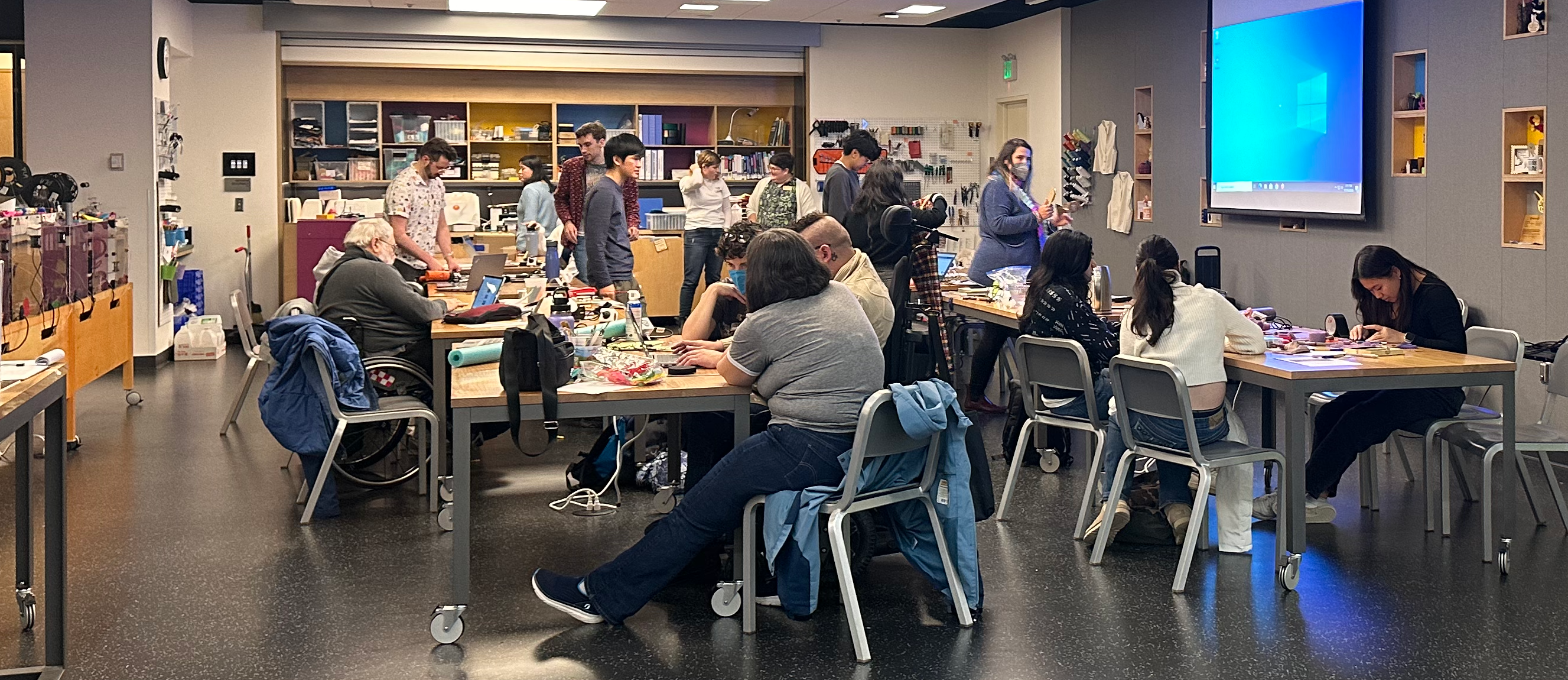 A well lit room at the Bellevue Library Makerspace. Rolling tables and flexible seating for accessibility, with several 3D printers on the left. Work tables are covered with projects in progress and surrounded by about 18 people.