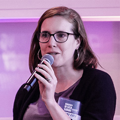 Amy Hurst, a white woman with brown hair and glasses, holding a microphone and talking in front of a large display screen that casts a purple light.