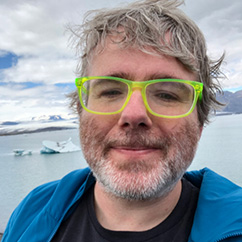 Shaun Kane has grey hair and a grey beard and bright green glasses. In this image he is posing in front of a frozen lake in Iceland.