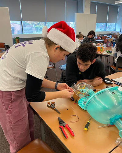 A female student wearing a Santa hat and a hearing aid, demonstrates how to add a switch to make a toy more usable by a child with a disability. She and the male student are focused on the detailed work.