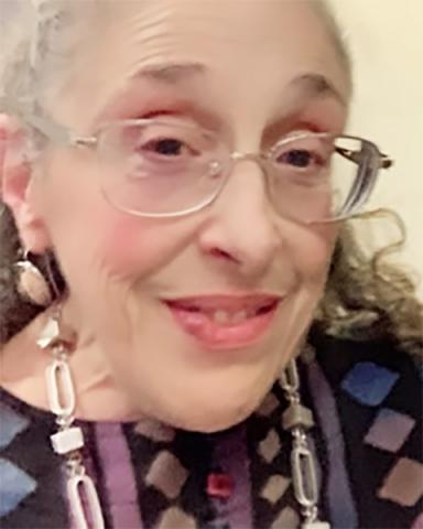 A close-up photo of Devva Kavnitz, a woman with gray hair pulled back, light skin, and rosy cheeks. She is wearing a colorful geometric design top with bold silver jewelry. 