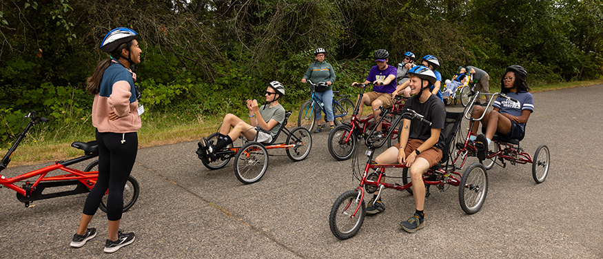 Nine people pausing on a variety of adaptive bicycles. One participant leads a discussion. They are on a wide, paved park trail.