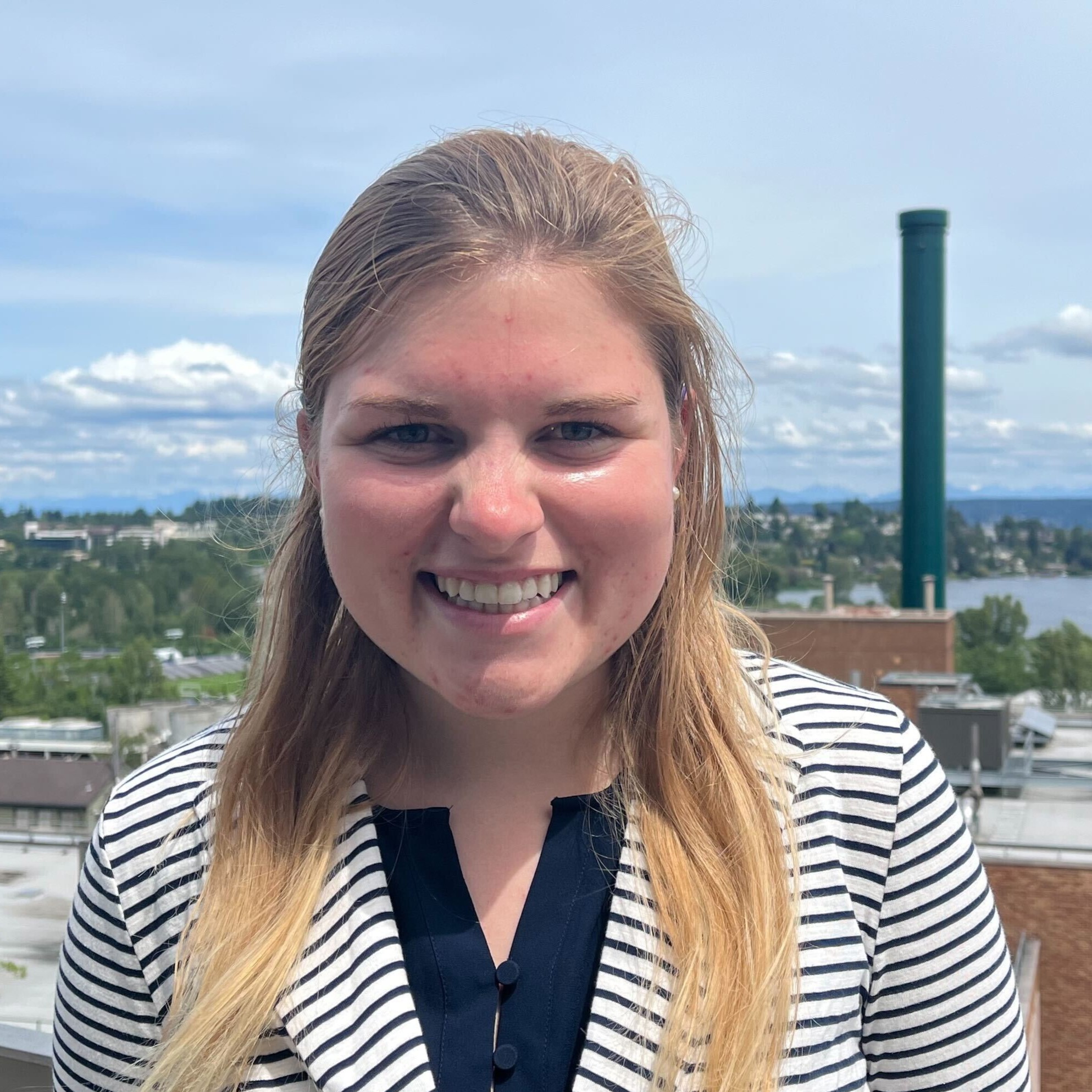 Mia Hoffman is smiling while standing on a sunny terrace on the UW Seattle campus. She has long blond hair and is wearing a striped jacket.