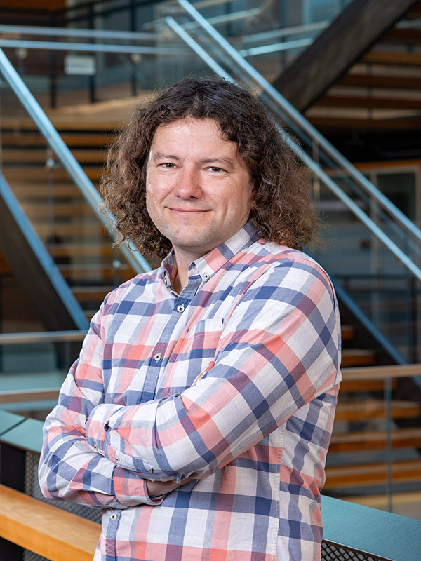 James Fogarty, a white man wearing a pink and blue plaid shirt with long, curly brown hair. He is posed with arms crossed and a warm smile.