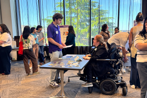 Kayla Brown seated in a power wheelchair UW DO-IT's table. She is talking to a community member holding a flyer from the table.