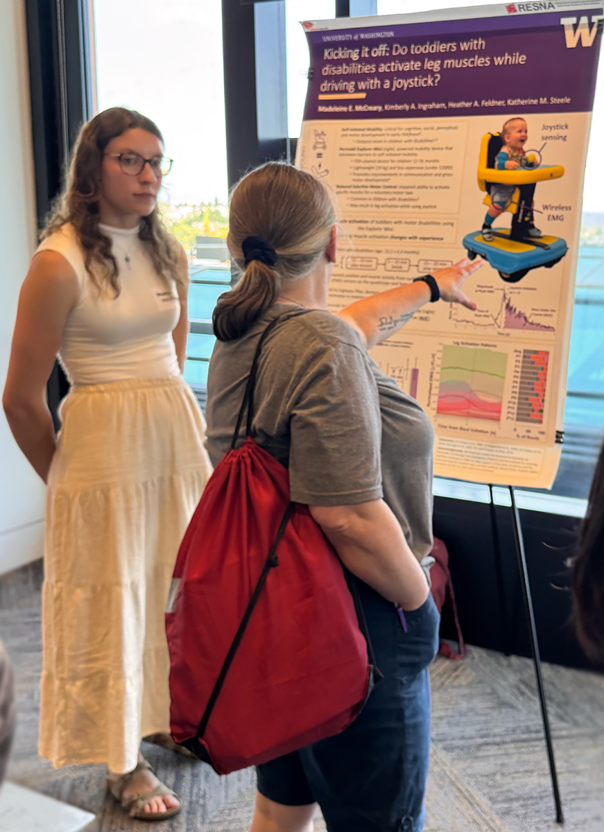 Madeleine McCreary fielding questions from a research showcase visitor who is gesturing toward McCreary's research poster, Do toddlers with disabilities activate leg muscles while driving with a joystick?