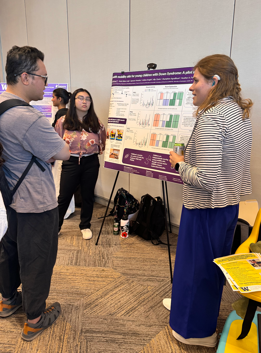 Mia Hoffman stands to the right of the research poster on mobility aids for children with Down Syndrome. She is in discussion with a community member and another student researcher.