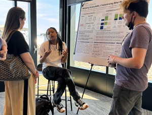 Aaleyah Lewis, seated on a stool next to the Modeling Accessibility research poster, talks to a community member and fellow student Jesse Martinez.