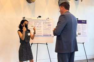 Aashaka Desai gestures with two hands and stands beside her research poster on Multilingual Captioning. A community member listens intently.