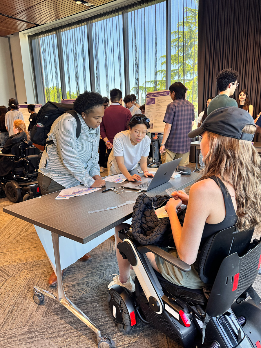 Two community members from Open Doors for Multicultural Families look at project information on a table and a laptop. The table is hosted by a young woman in a power wheelchair.