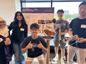 Tamanna Motahar and stduent researchers pose in front of their research poster on a robotic screen positioning tool. Two students hold metal prototypes and an earlier cardboard prototype is visible.