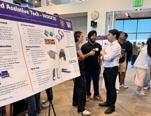 A very large research poster from WOOF3D is in the foreground. Three community members and researchers in discussion are in the background.