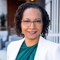 Michele Williams, a Black woman with natural locs. She wears a teal top under a white blazer and clear-rimmed glasses.