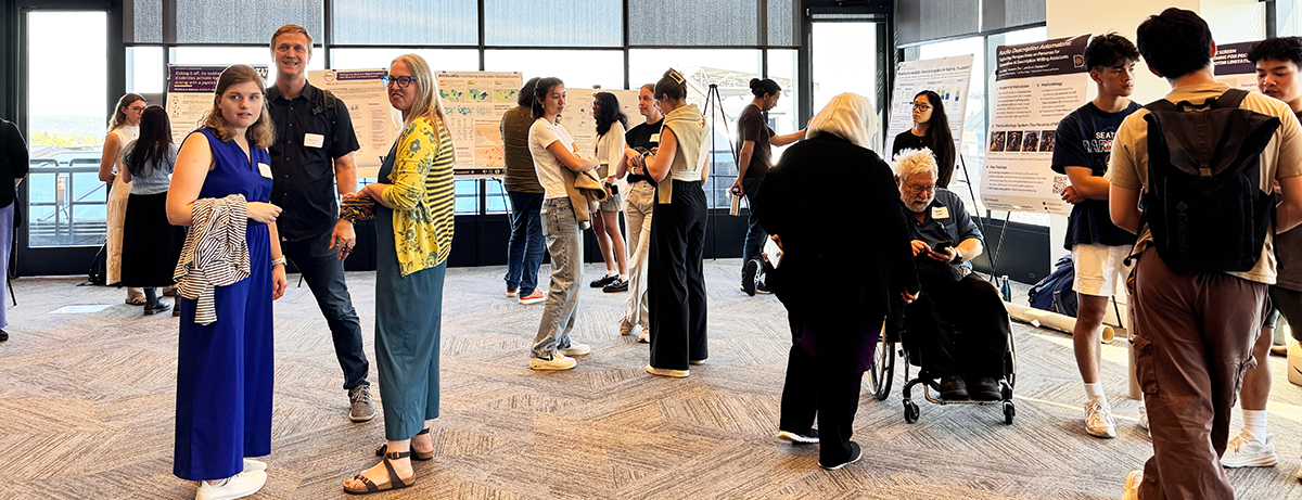 CREATE Research Showcase 2025 attendees gather in discussions around research posters. To the left, HuskyADAPT student lead Mia Hoffman, CREATE Education Director Mark Harniss, and CREATE associate director Heather Feldner stand together, looking toward the camera.