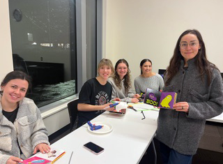 Four students sit at a work table with supplies for adapting books. Sloane stands in front, holding an adapted children’s book.