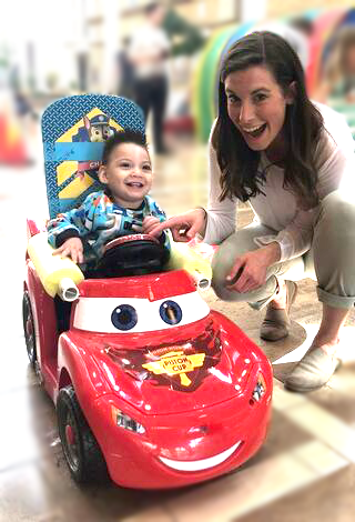Bethany Sloane, a white woman with long, brown hair, smiles brightly while crouching next to a red ride-on car occupied by a young child smiling just as brightly.