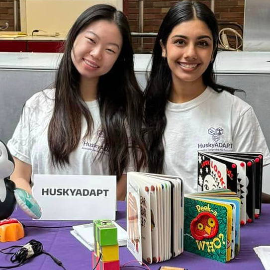 Two HuskyADAPT students, both women with long, brown hair, sit at an event table displaying adapted books with rhinestones helping separate pages.