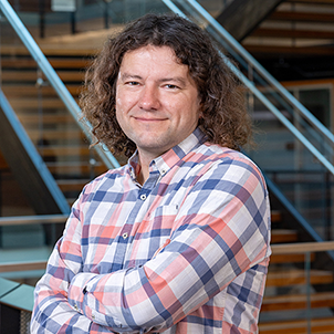 James Fogarty, a white man wearing a pink and blue plaid shirt with long, curly brown hair. He is posed with arms crossed and a warm smile.