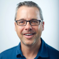 Jonathan Warren, a white man with salt and pepper hair and dark-rimmed eyeglasses. He wears a blue button-down shirt and smiles happily.