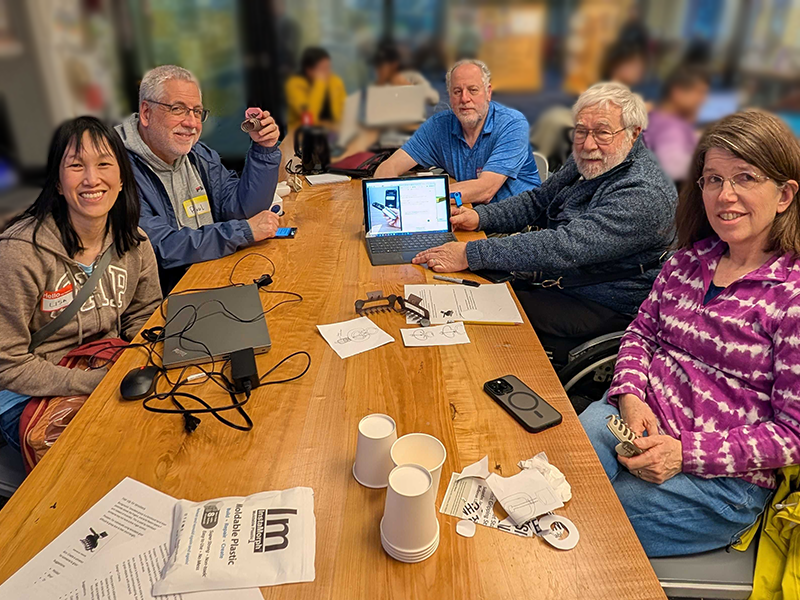 Five adults seated at a work table. They hold various large butterfly/ claw hairclips and the table holds laptops and prototyping marterials.