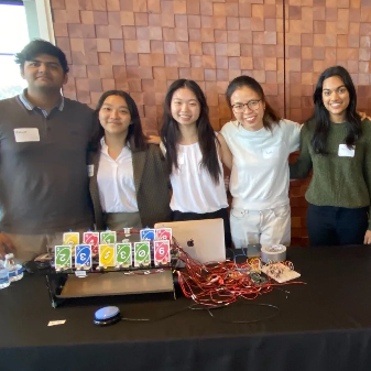Five students stand at a demonstration table with a game where playing cards are hooked to wires.