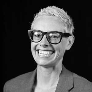Black and white headshot of Anna Ziverts. They smile effusively and have close-cropped platinum hair and large black-framed eyeglasses.