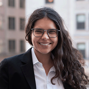Ariana Aboulafia, smiling outside in front of a city background. With long dark hair, wearing a white collared shirt and black jacket.